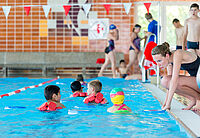 Kinder werden im Schwimmbecken zum Schwimmen angewiesen, Trainerin kniet am Beckenrand