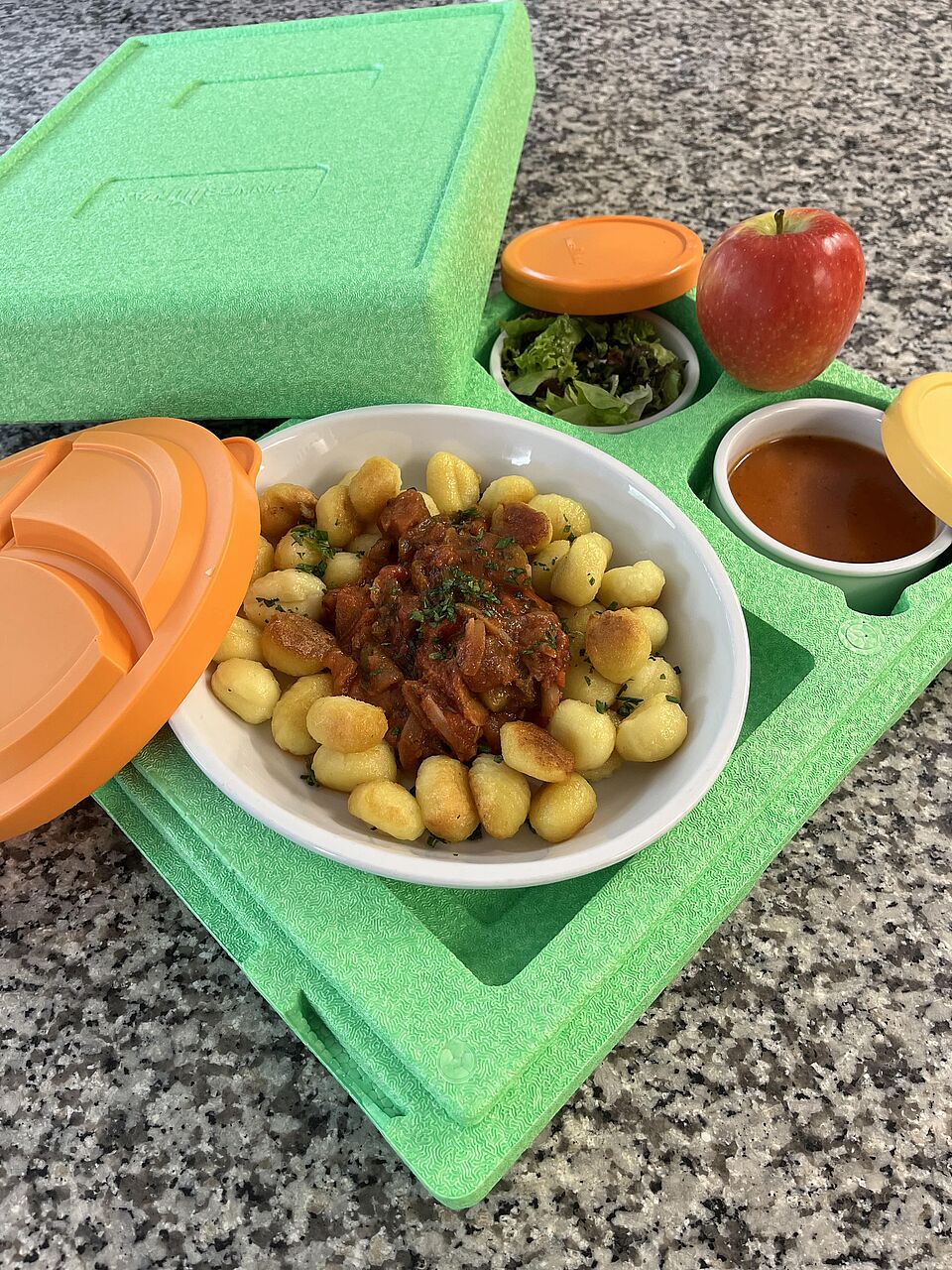 Menü für Essen auf Rädern: ein großer Teller mit Gnocchi und Tomatensoße, dahinter Schale mit Suppe und Schale mit Salat, alles in einer Wärmebox bereitgestellt