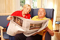 Eine DRK Mitarbeiterin liest einem Bewohner die Zeitung vor. Der Bwohner und die Mitarbeiterin halten zusammen die Zeitung.