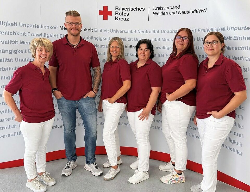 BRK_Tagespflege_Eschenbach__Gruppenfoto Auf dem Bild sind sechs Mitarbeiter*innen aus der Tagespflege zu sehen. Sie tragen eine weiße Jeans und ein weinrotes T-Shirt - stehend vor der Fotowand