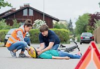 Erste Hilfe am Fahrradfahrer auf der Straße, Helmabnahme von den beiden Ersteintreffern, Frau trägt Warnweste