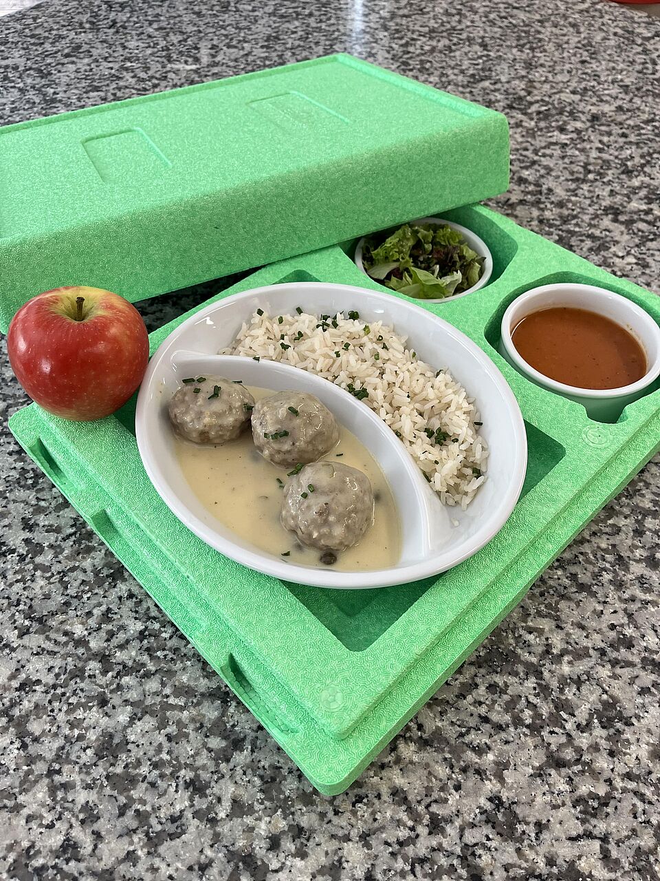 Menü für Essen auf Rädern: ein großer Teller, aufgeteilt in 2 Hälften, Königberger Klöpse mit Reis, dahinter Schale mit Suppe und Schale mit Salat, alles in einer Wärmebox bereitgestellt