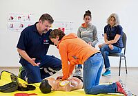 Erste Hilfe Kurs mit HLW Training, Ausbilder erklärt Teilnehmerin die HLW, im Hintergrund sitzen zwei weitere Teilnehmer