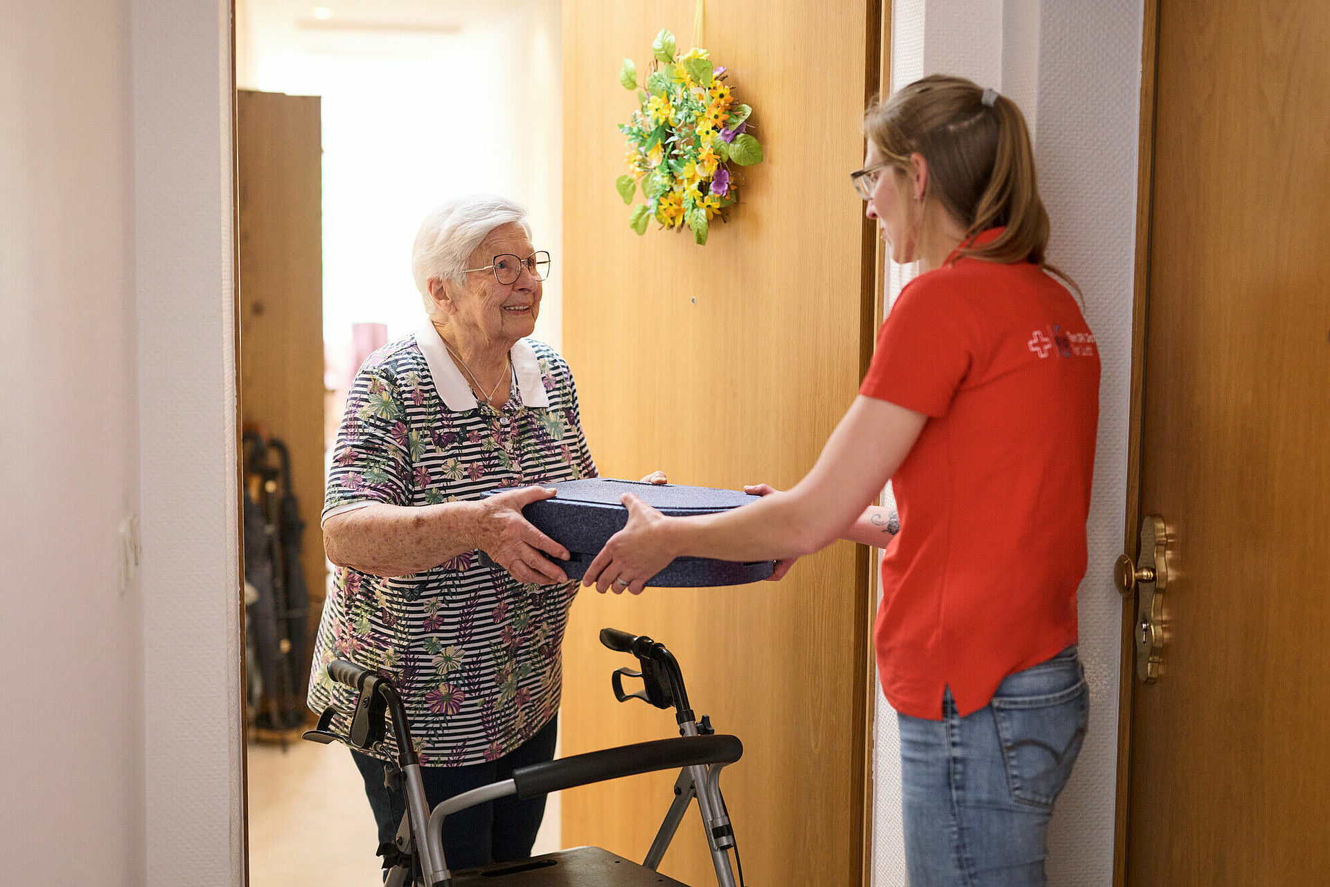 DRK Mitarbeiterin übergibt Essen an ältere Dame, Essen befindet sich in einer Warmhaltebox, Übergabe findet an der Haustüre statt
