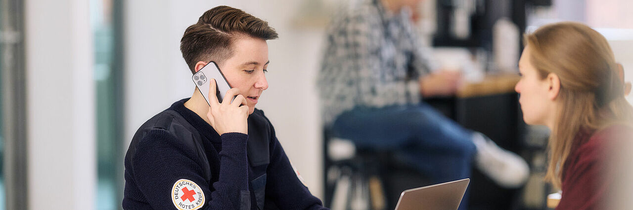 BRK Mitarbeiterin telefoniert am Handy, sie symbilisiert die Ansprechpartnerin für Anfragen, gegenüber sietzt eine Interessentin