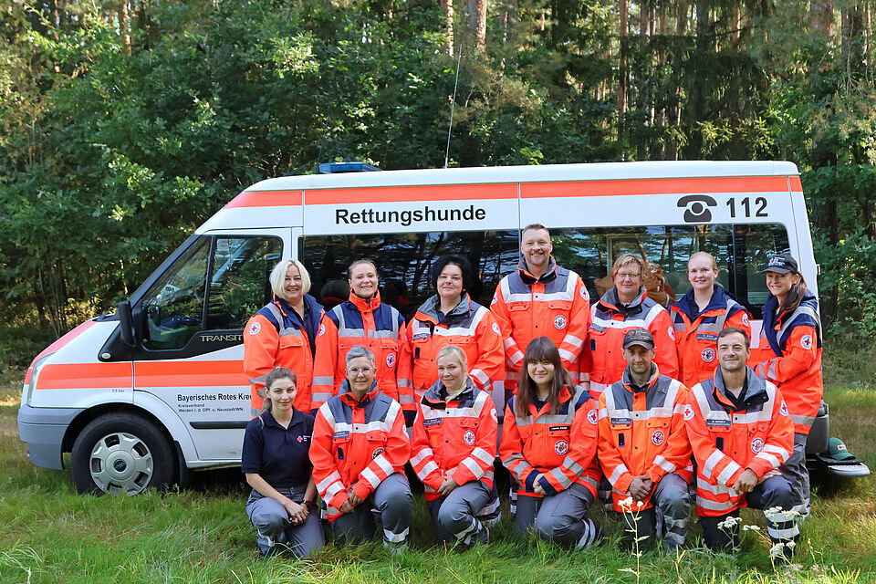 Einsatzfahrzeug BRK Rettungshundestaffel, davor stehend 13 ehrenamtliche BRK Mitglieder in Einsatzkleidung, rote Jacke