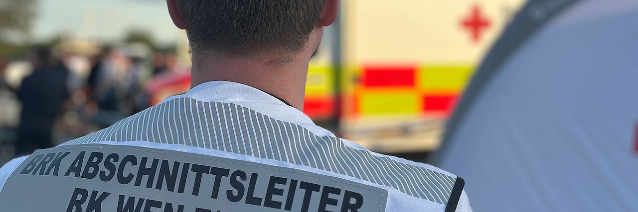 Sanitätswachdienst Abschnittsleiter mit weißer Weste, Hintergrund RTW und Veranstaltung
